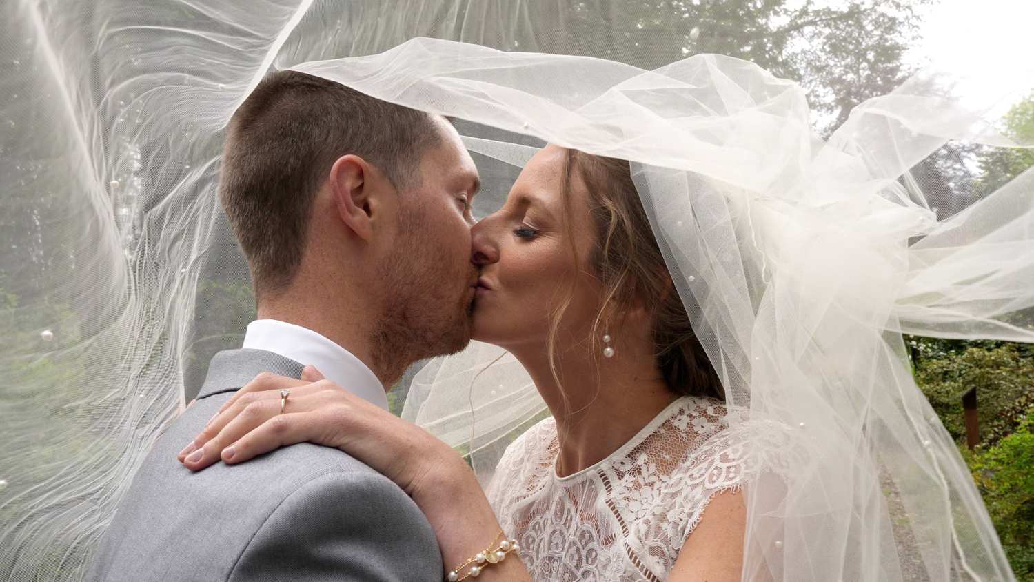 Romantic wedding couple shares tender kisses under a flowing white veil in an outdoor garden setting.