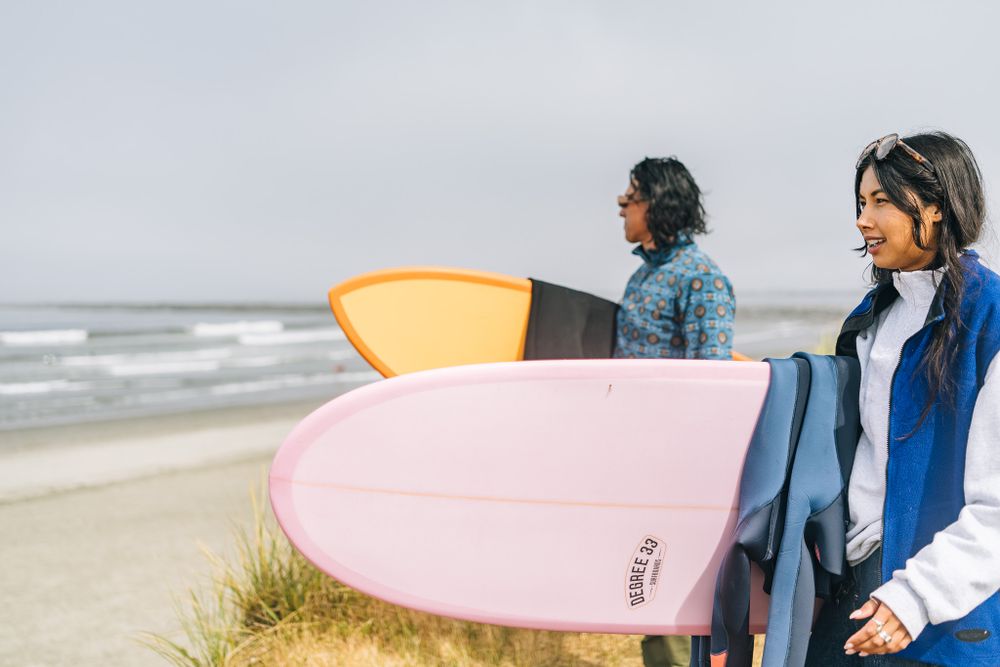 Two surfers holding pink and orange surfboards stand on a beach looking out at the ocean on a cloudy day.