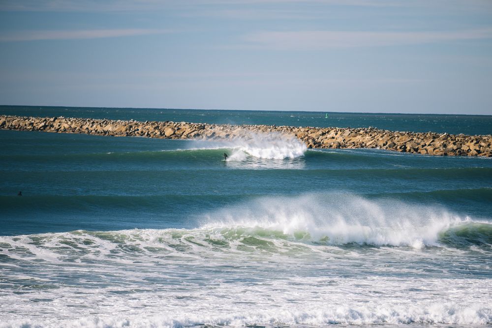 Ocean waves break near a rocky jetty on a clear day with blue skies and white surf.