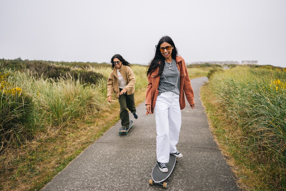 Two people skateboarding on a paved path surrounded by tall grass and yellow wildflowers on a sunny day.