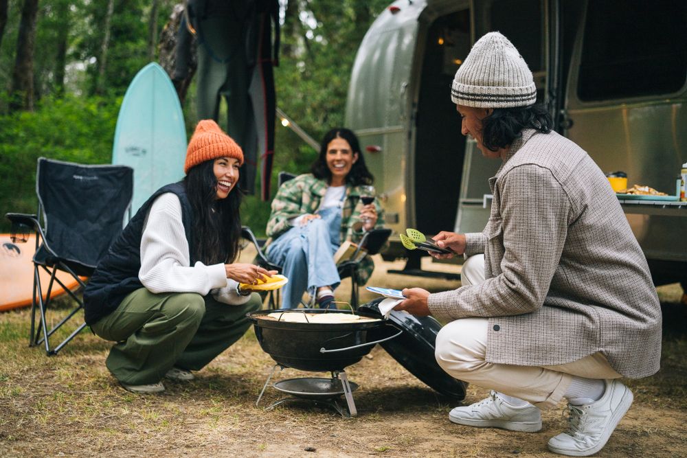 Friends camping and cooking outdoors together around a portable grill with surfboards and an Airstream trailer in background.