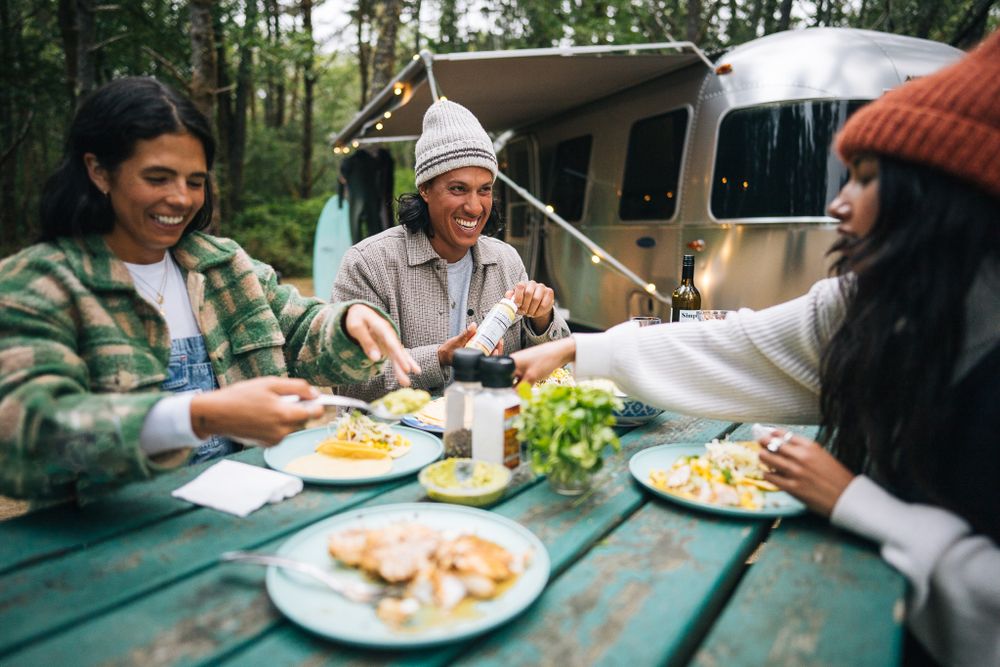Friends enjoying an outdoor meal together at a picnic table near a camping van in the forest.