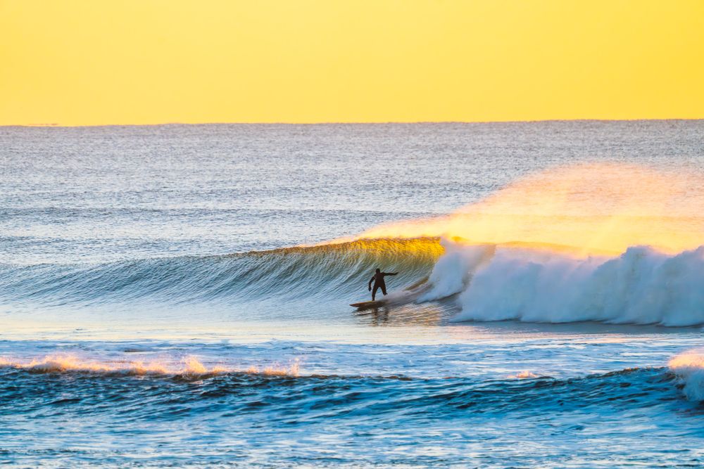 A surfer rides a breaking wave during golden sunset with vibrant yellow sky and ocean spray.