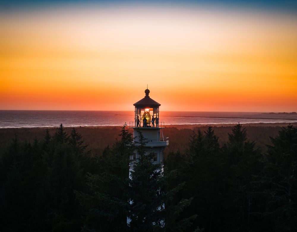 Lighthouse silhouetted against vibrant orange sunset sky with evergreen forest in foreground.