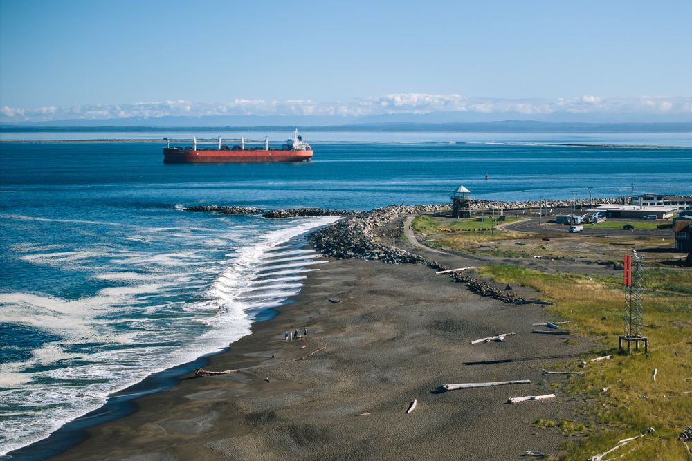 A cargo ship sails past a rugged coastline with black sand beach, waves, and rocky shoreline in Hokkaido, Japan.