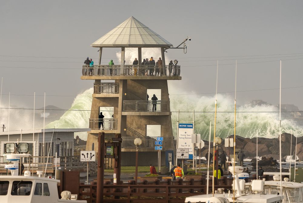 Large waves crash near a wooden lighthouse observation tower at a harbor while spectators watch from above.
