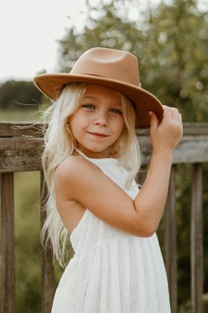 Sunlit portrait with a brown felt hat and white summer dress against a wooden fence.