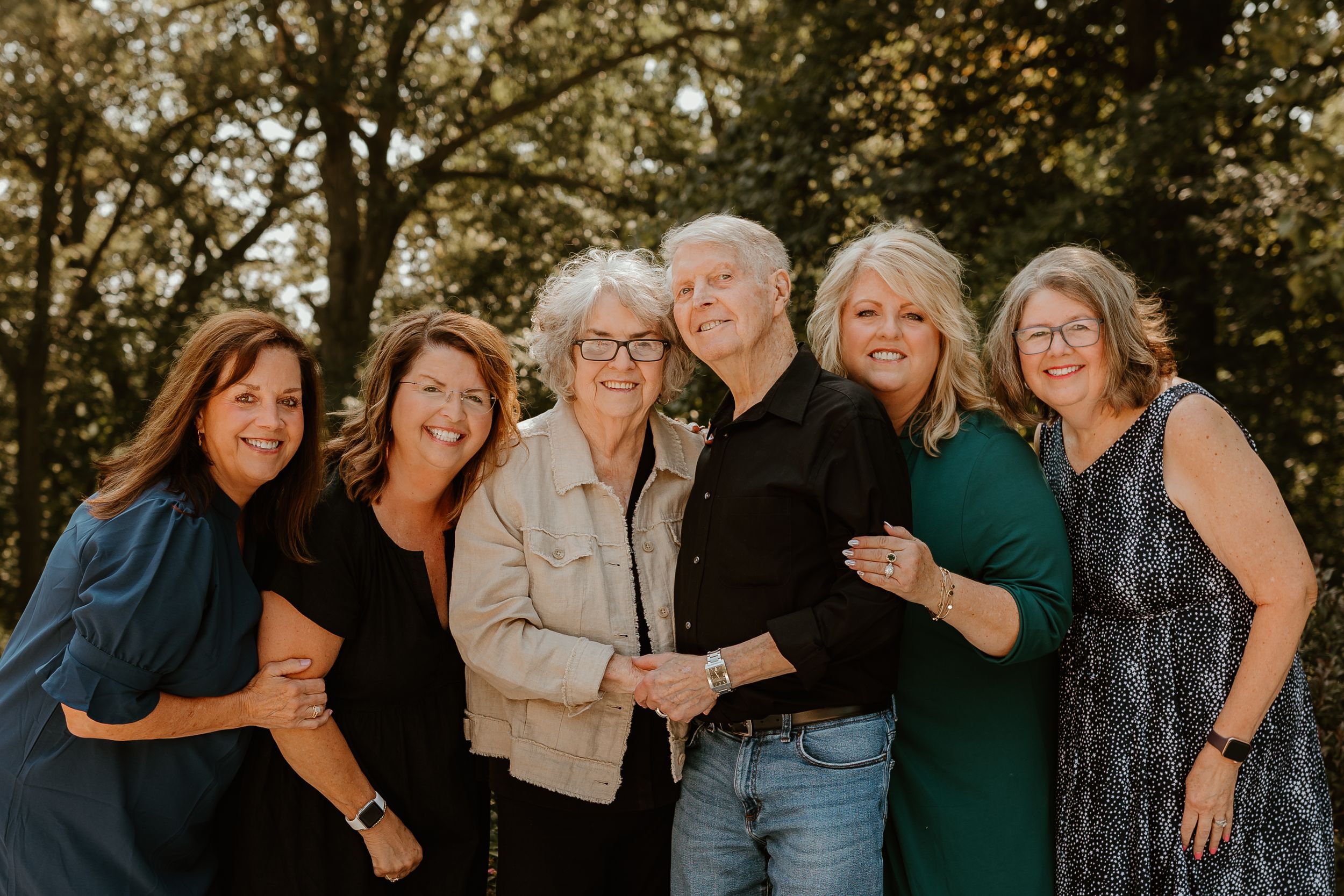 A happy multi-generational family poses together outdoors in warm sunlight against a backdrop of trees.