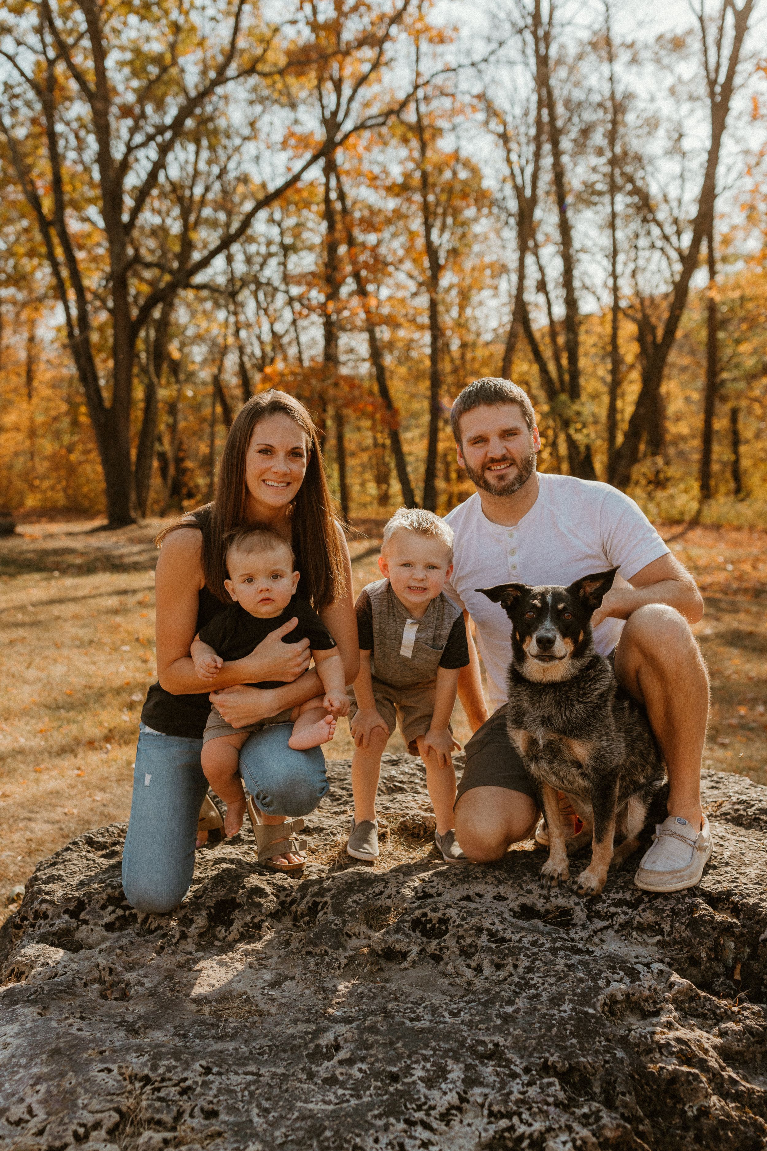 A family portrait with their dog sitting on a rock in a fall forest setting.