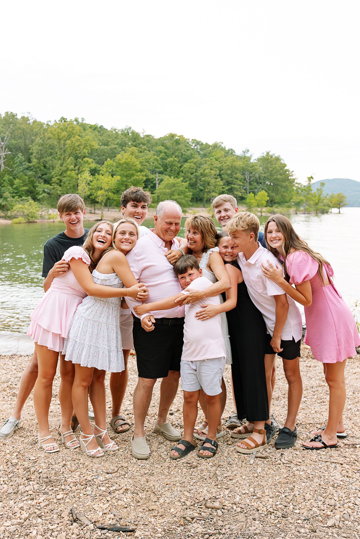 Grandchildren and grandparents hugging and laughing together on beach with lake and mountains in background.