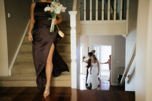 Bridesmaids in elegant dark gowns descend a grand staircase, one holding a white bouquet, as a bride prepares nearby.