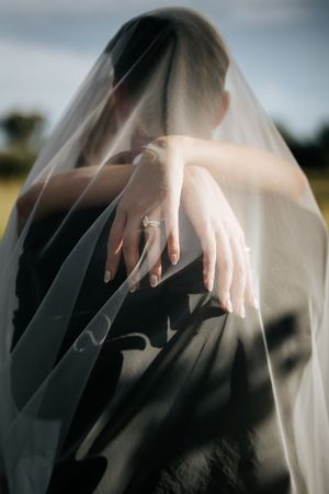 Bride's hands showing diamond engagement ring draped under sheer wedding veil in soft natural light.