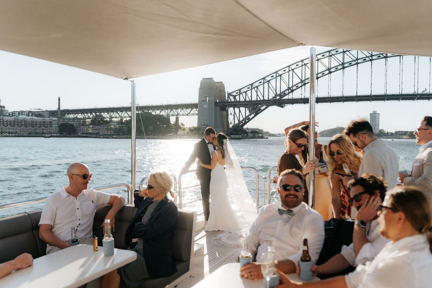 Wedding couple kissing on a luxury boat with Sydney Harbour Bridge backdrop, surrounded by guests celebrating.