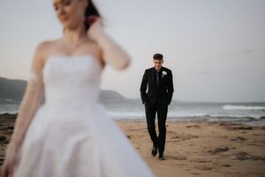 A groom in a black suit walks toward the bride in a white strapless gown on a sandy beach at dusk.