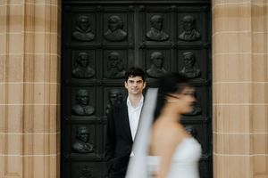 Groom in black suit stands before ornate dark bronze doors while bride in white veil walks past in blur.