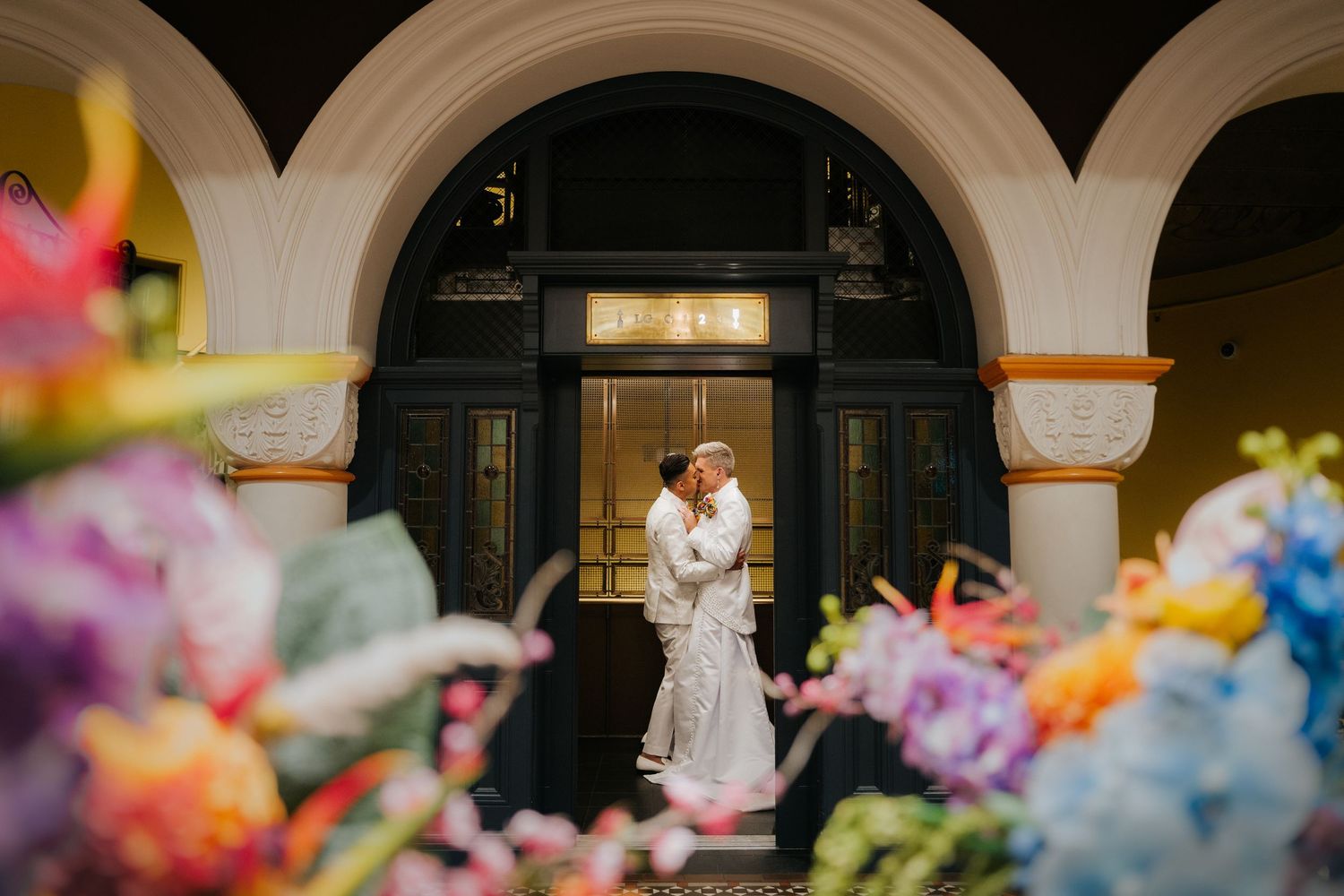 Bride and groom embracing in a grand arched doorway surrounded by colorful floral arrangements.