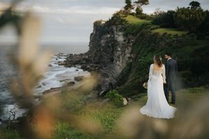 Bride and groom standing on dramatic coastal cliff overlooking ocean waves at golden hour.