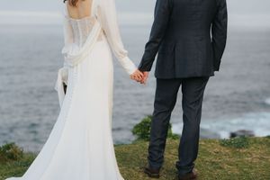 Newlyweds holding hands facing the ocean in elegant white wedding dress and charcoal suit.