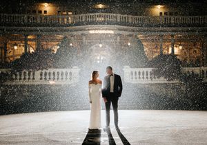 Romantic couple in wedding attire standing in falling snow outside illuminated grand building at night.