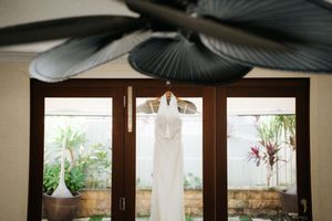 White wedding dress hanging on wooden French doors with tropical garden view and ceiling fan above.