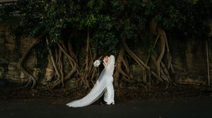 Bride in white gown kissing groom beneath dramatic twisted tree roots against a stone wall.