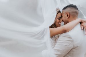 Bride and groom embrace tenderly under a flowing white veil in a romantic wedding portrait.