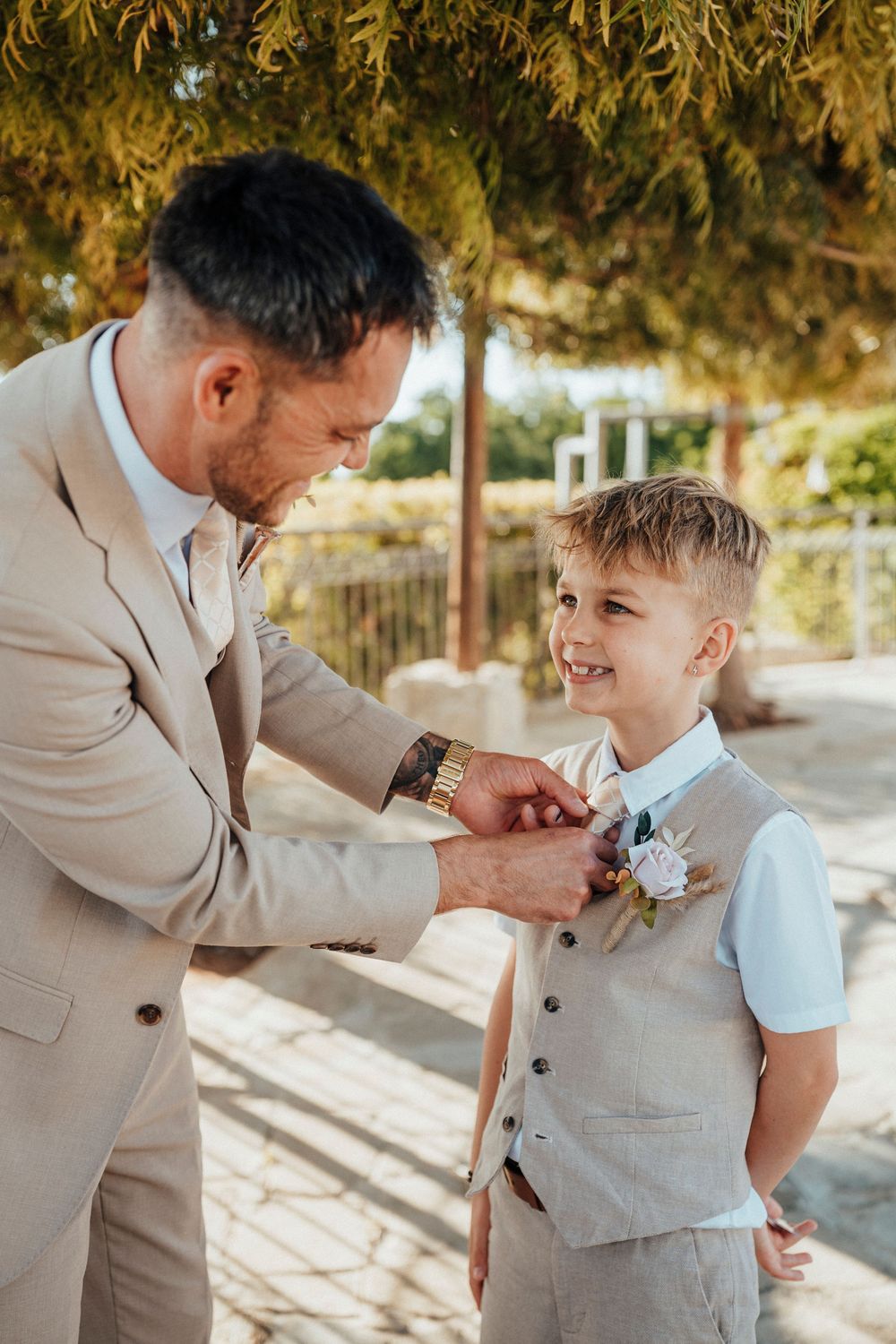 A tender moment as a person in a beige suit helps a young child with a light blue shirt adjust a boutonniere in sunlit setting.