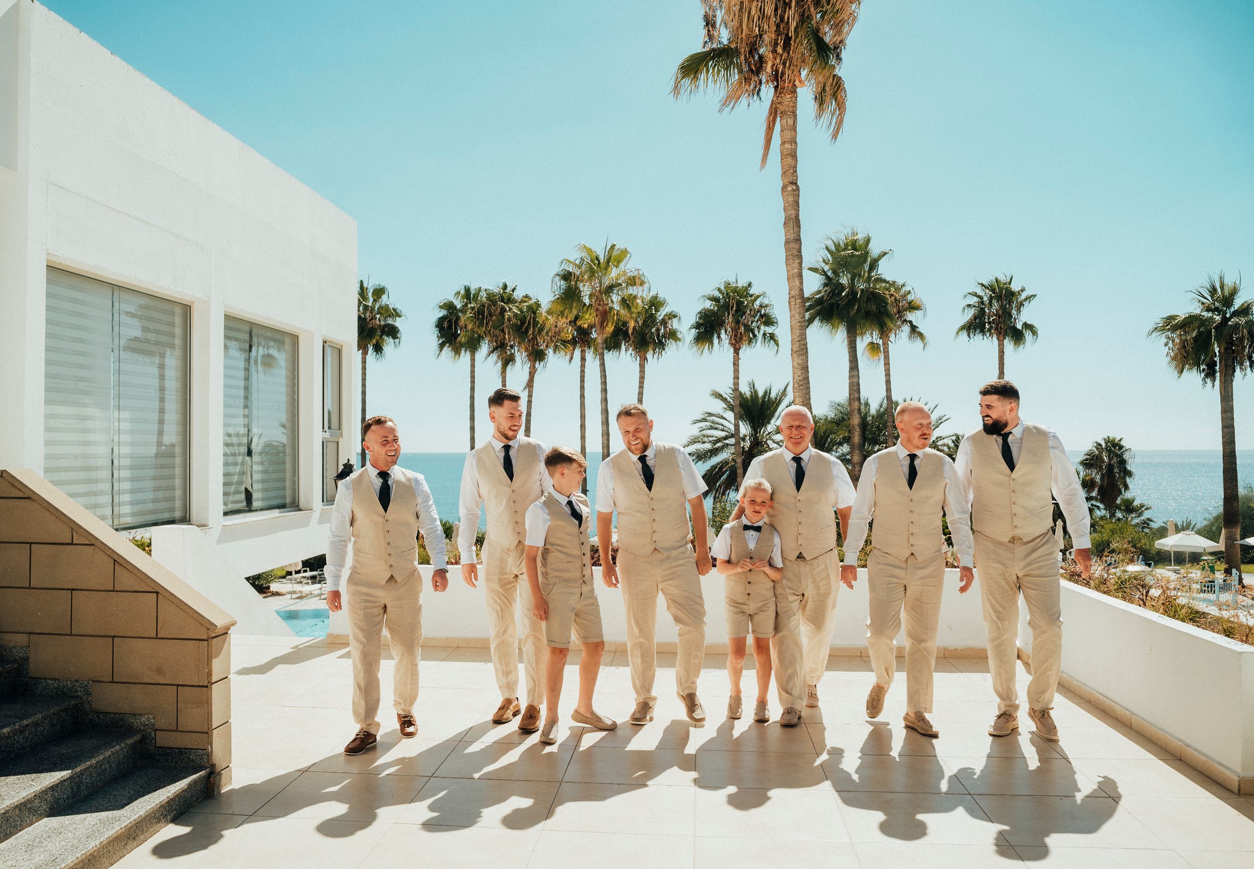 A group of people in beige suits cast long shadows while posing against a white wall with palm trees in the background.