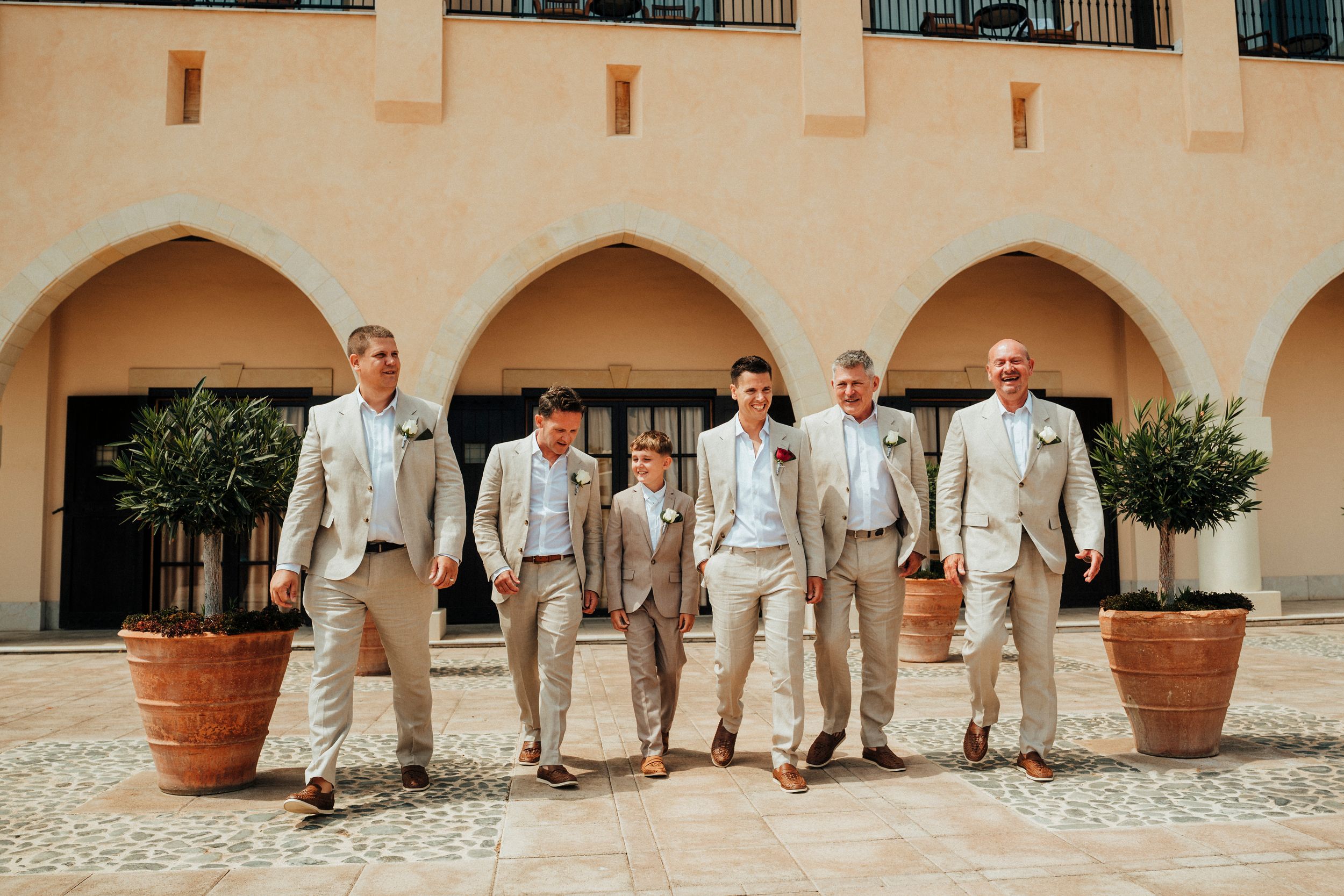 A group of groomsmen in light beige suits pose together against a Mediterranean-style building with arches.