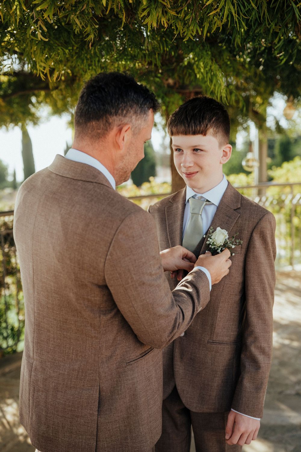 A tender moment captured between two people in brown suits adjusting boutonnieres outdoors under trees.