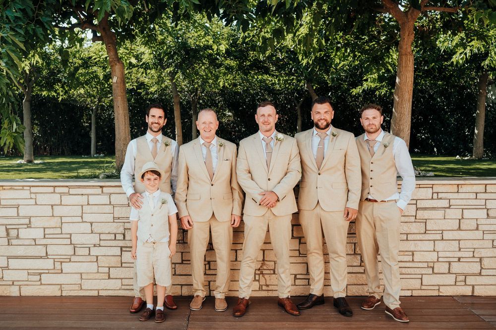 Wedding party in light colored attire poses casually against a white brick wall outdoors.