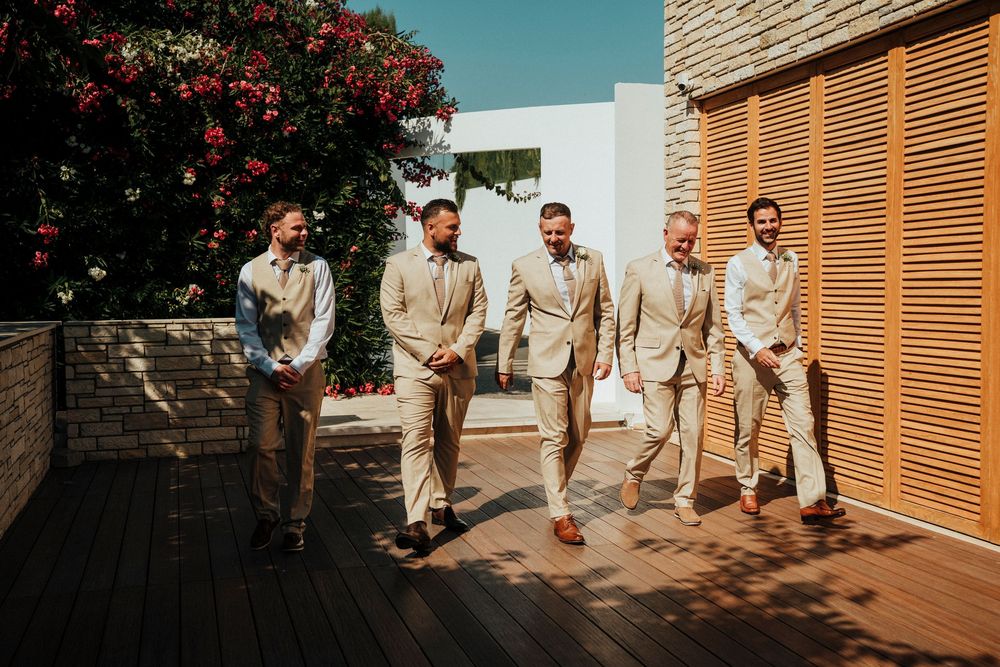 Groomsmen in beige suits walk together outside a modern building with red flowers in the background.