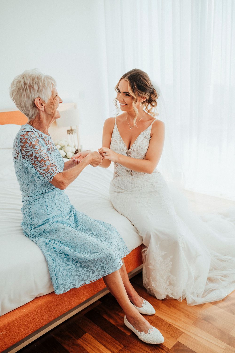 Two people share a tender moment while sitting on a wooden bench in a bright white room, one wearing a blue dress.