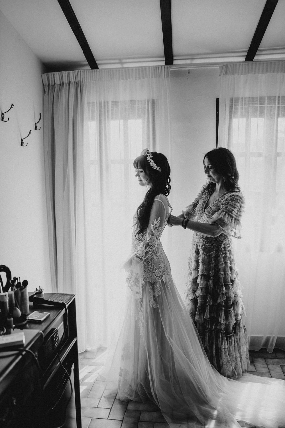 A series of black and white photos showing a bride getting ready with assistance in a bright room with sheer curtains.