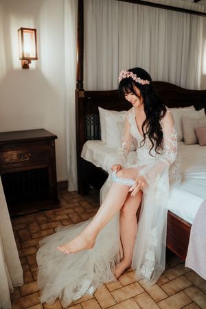 A bride in a flowing white dress sits on a bed while putting on her wedding garter in a rustic room.