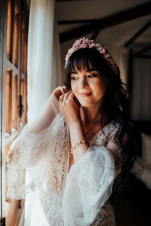 A bride in a delicate white lace robe adjusts her earrings while getting ready near a window.