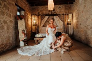 A bride in a white lace wedding dress adjusts her shoe while sitting in a rustic stone room with warm lighting.