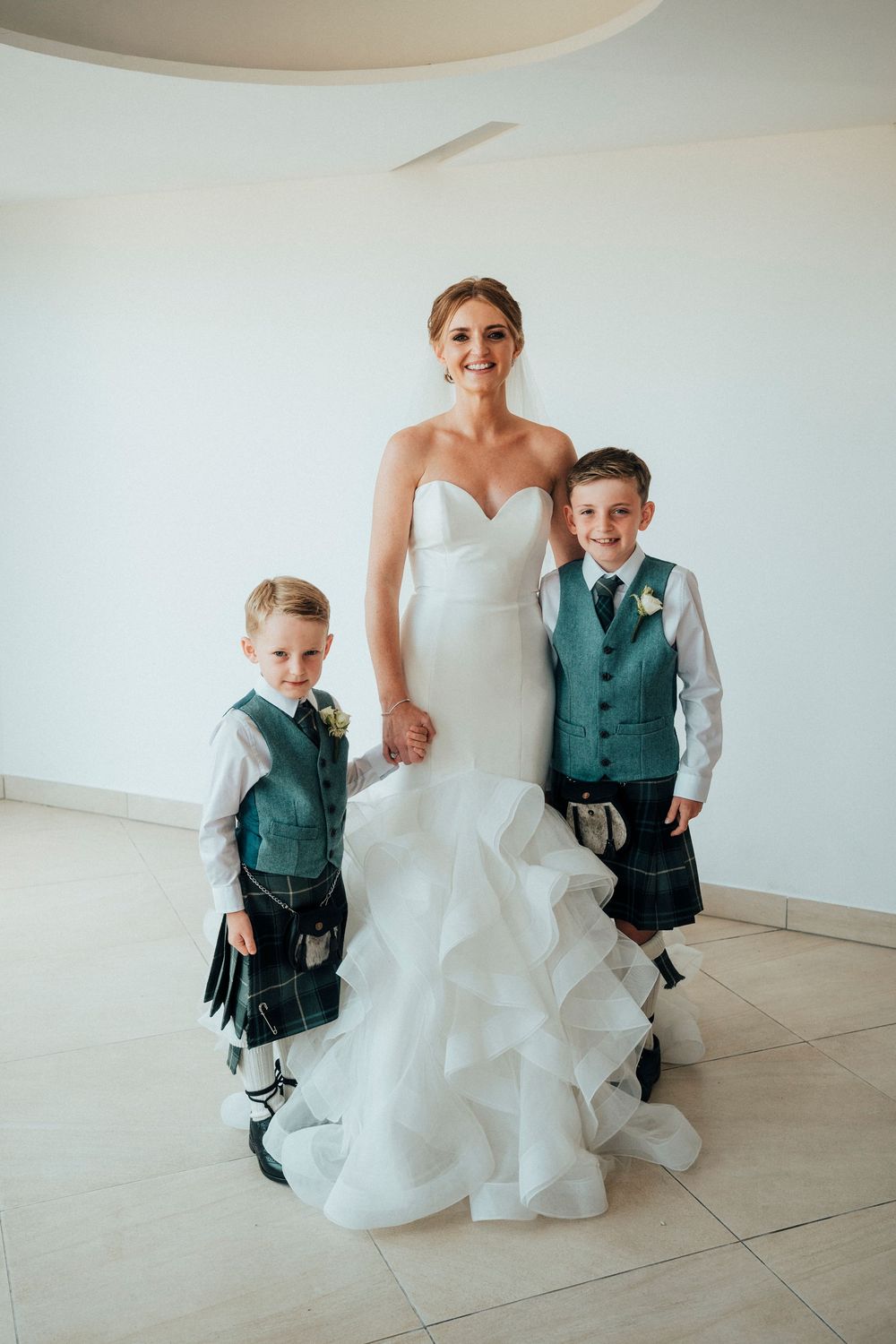 A bride in a white ruffled gown kneels beside a young boy in a teal vest and kilt at a wedding celebration.