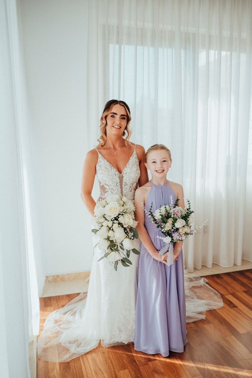 Wedding party in lavender dresses and white gown pose together in an elegant indoor setting with sheer curtains.