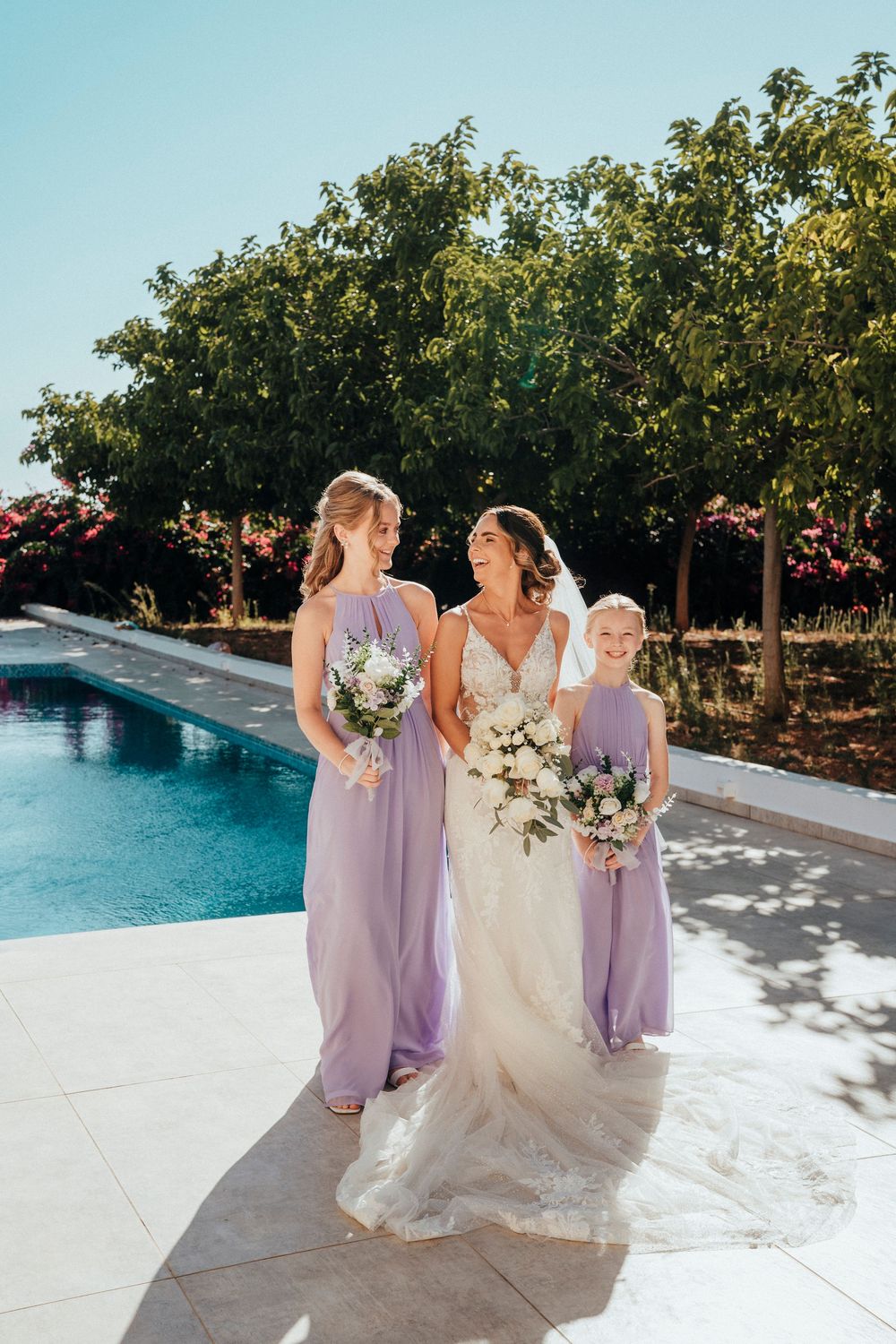 Wedding party in lavender dresses and white gown pose by a poolside surrounded by trees on a sunny day.