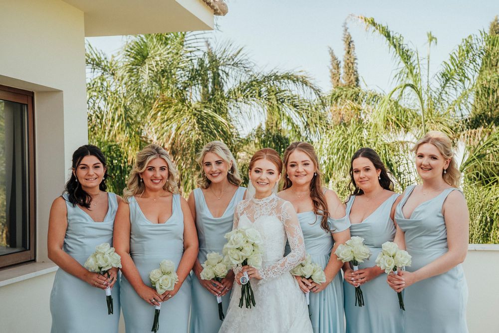 Bridesmaids in light blue dresses pose together with white bouquets at an outdoor venue with palm trees.