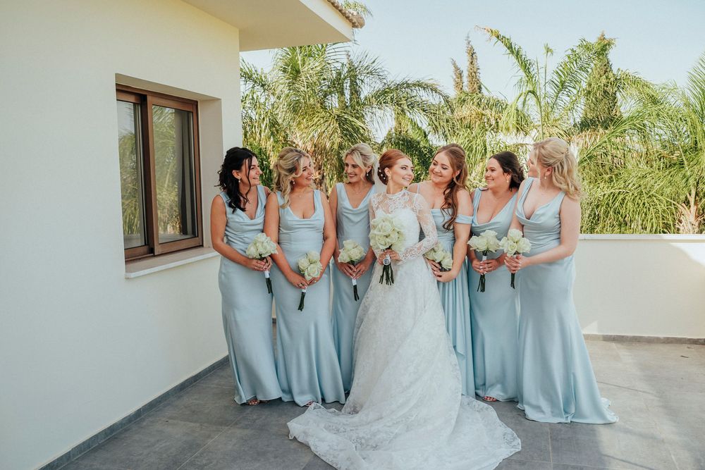 Wedding party in light blue bridesmaid dresses poses with bride in white gown on sunny outdoor terrace.