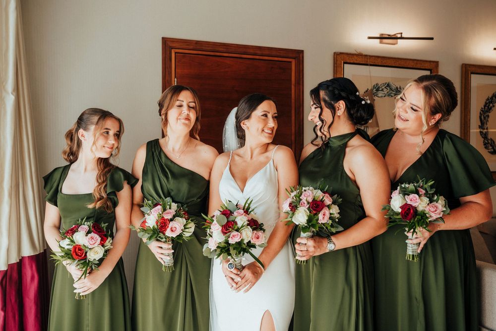 A group wearing matching green dresses holds pink and white bouquets while standing in a line indoors.