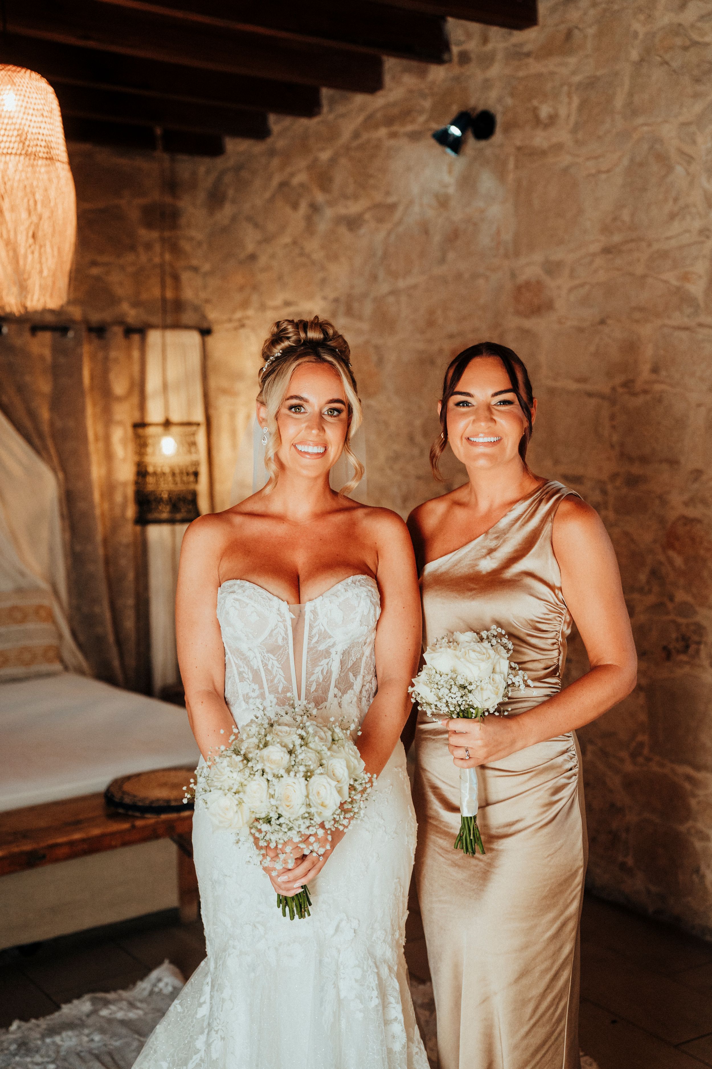 Wedding party members in champagne and white dresses hold delicate baby's breath bouquets in rustic stone venue.