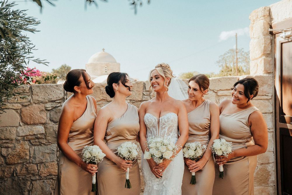 A group of bridesmaids in matching gray dresses laugh together while posing against a rustic stone wall at sunset.