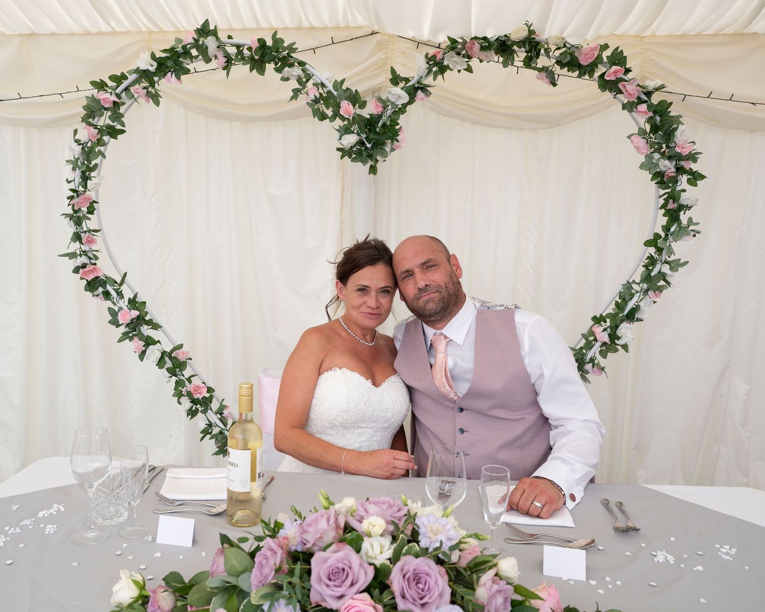 A wedding couple poses at their reception under a floral heart-shaped arch with pink roses on the table.