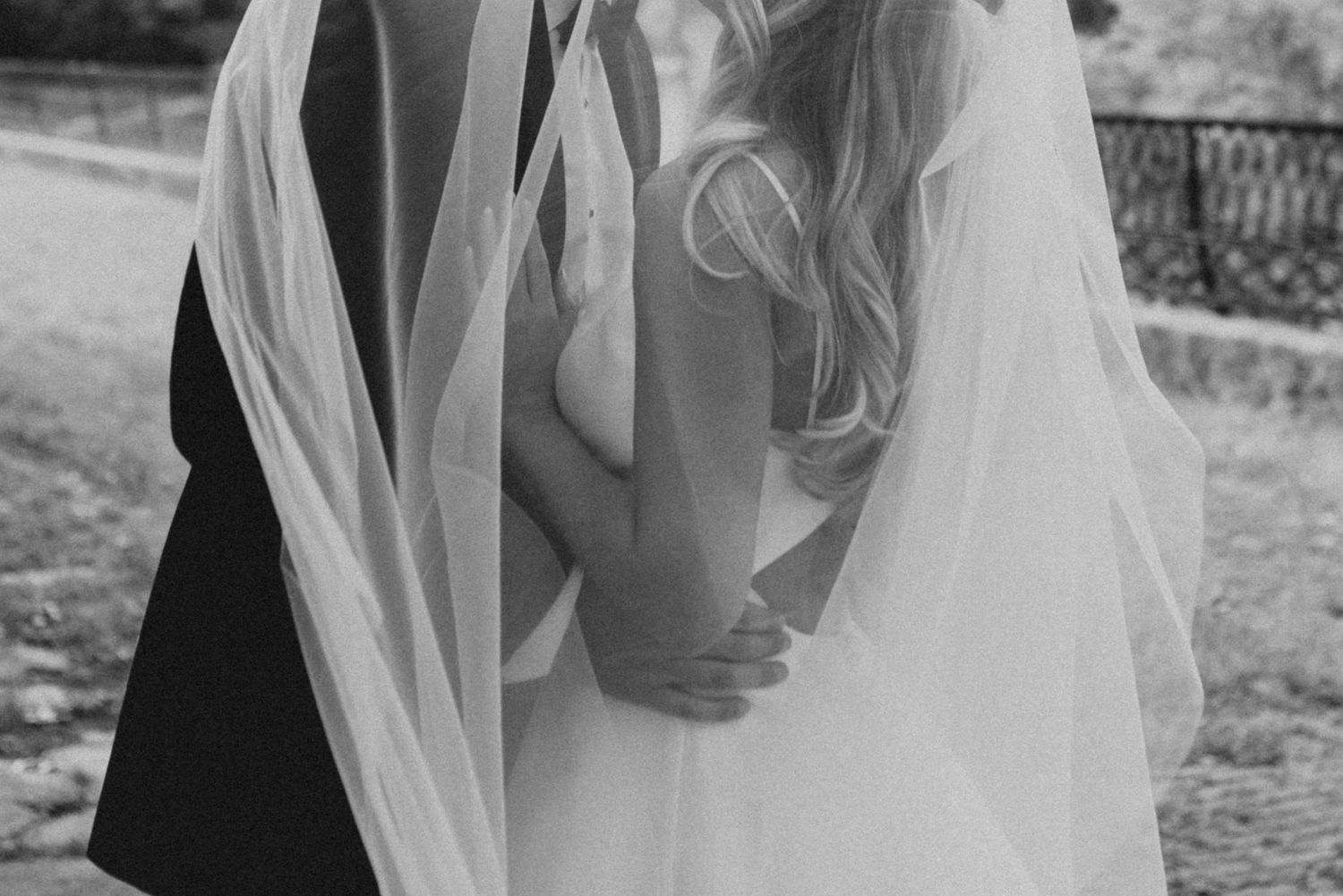 Romantic black and white photo of newlyweds embracing under a flowing veil outdoors.