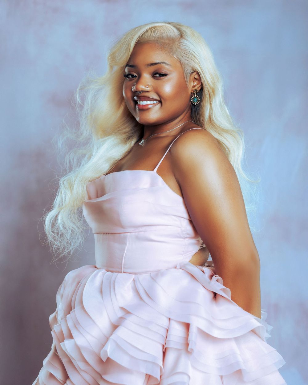 Young woman in pink ruffled dress with blonde wavy hair posing confidently against blue backdrop.