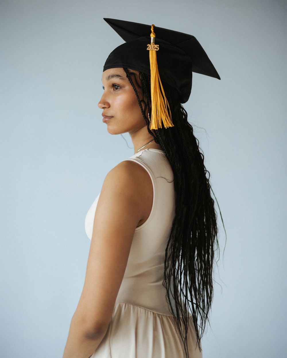 Graduate in cream dress and black mortarboard cap with gold tassel poses in profile against light gray background.