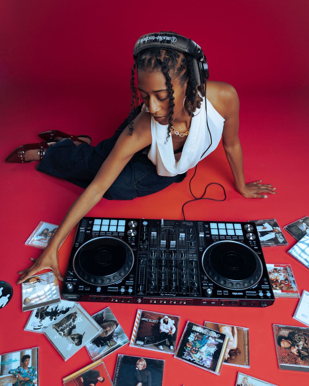 A young woman DJ sits on a red floor with headphones, a DJ controller, and scattered CD cases around her.